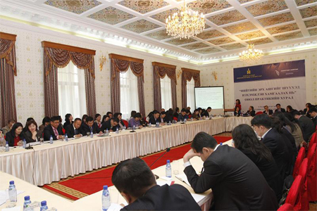 A conference in Mongolia depicts an ornate room. Tables with satin white tablecloths circle the room and people wear suits around the rectangle. A poster hangs on the wall.