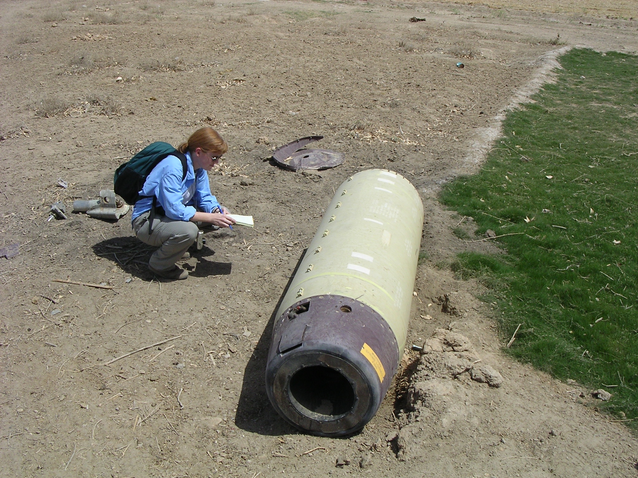 Bonnie examines a cluster munition in the dirt in Iraq. She bends down with a piece of paper.