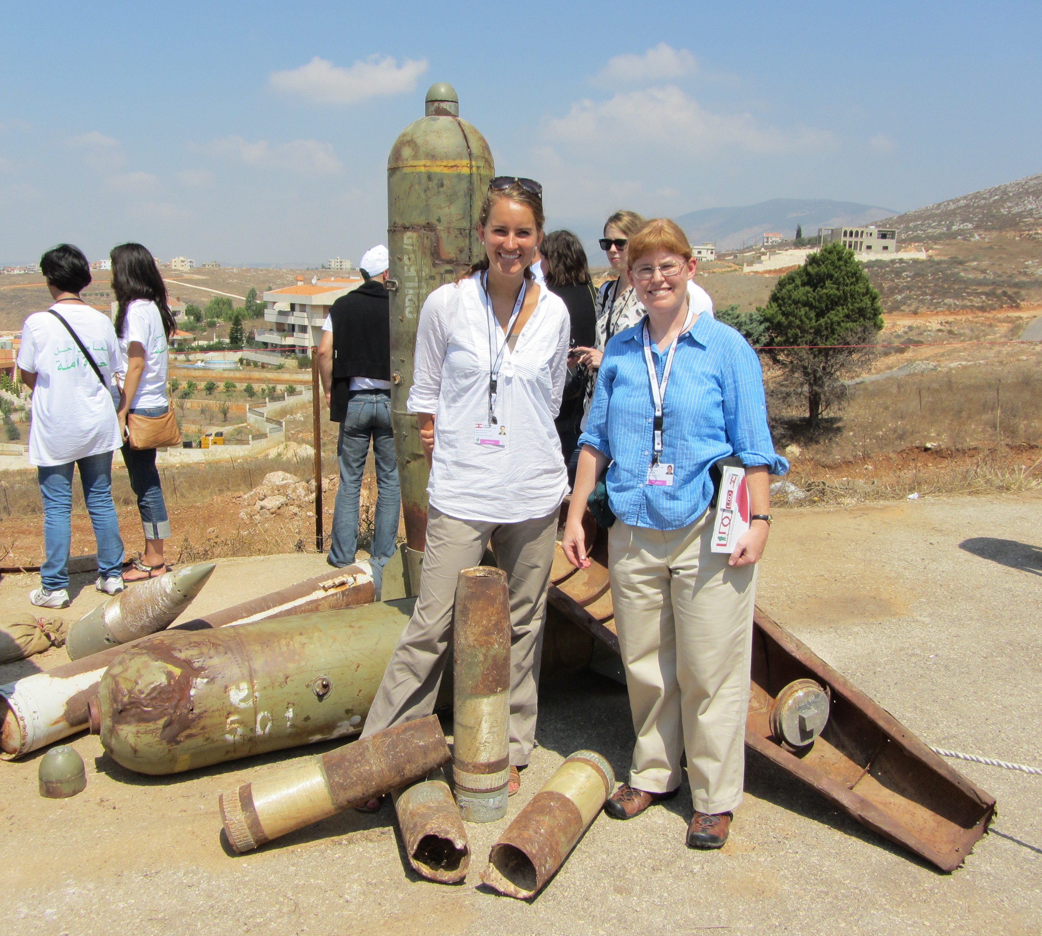 Two women stand smiling in a hot desert landscape; they stand over large rusty shells that look like weapons.