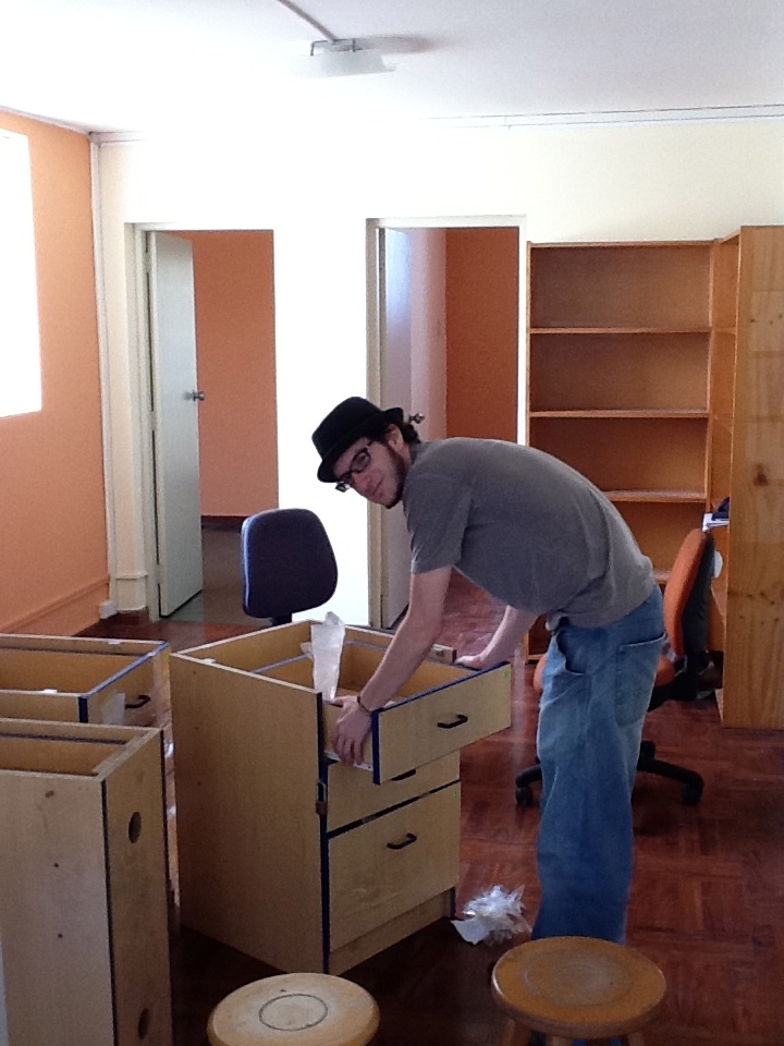 A man wearing a brimmed hat crouches over a set of drawers. He stands in front of boxes and empty bookshelves.