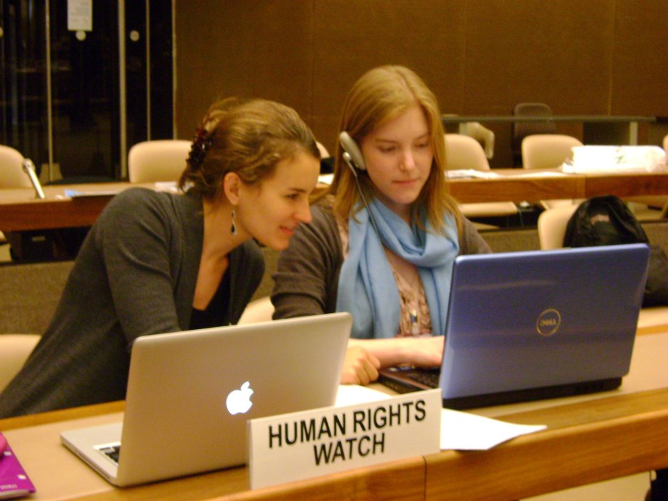 Nicolette Boehland leans in over Anna Crowe's shoulder behind two computers. There is a tent card in front of them that reads "Human Rights Watch."