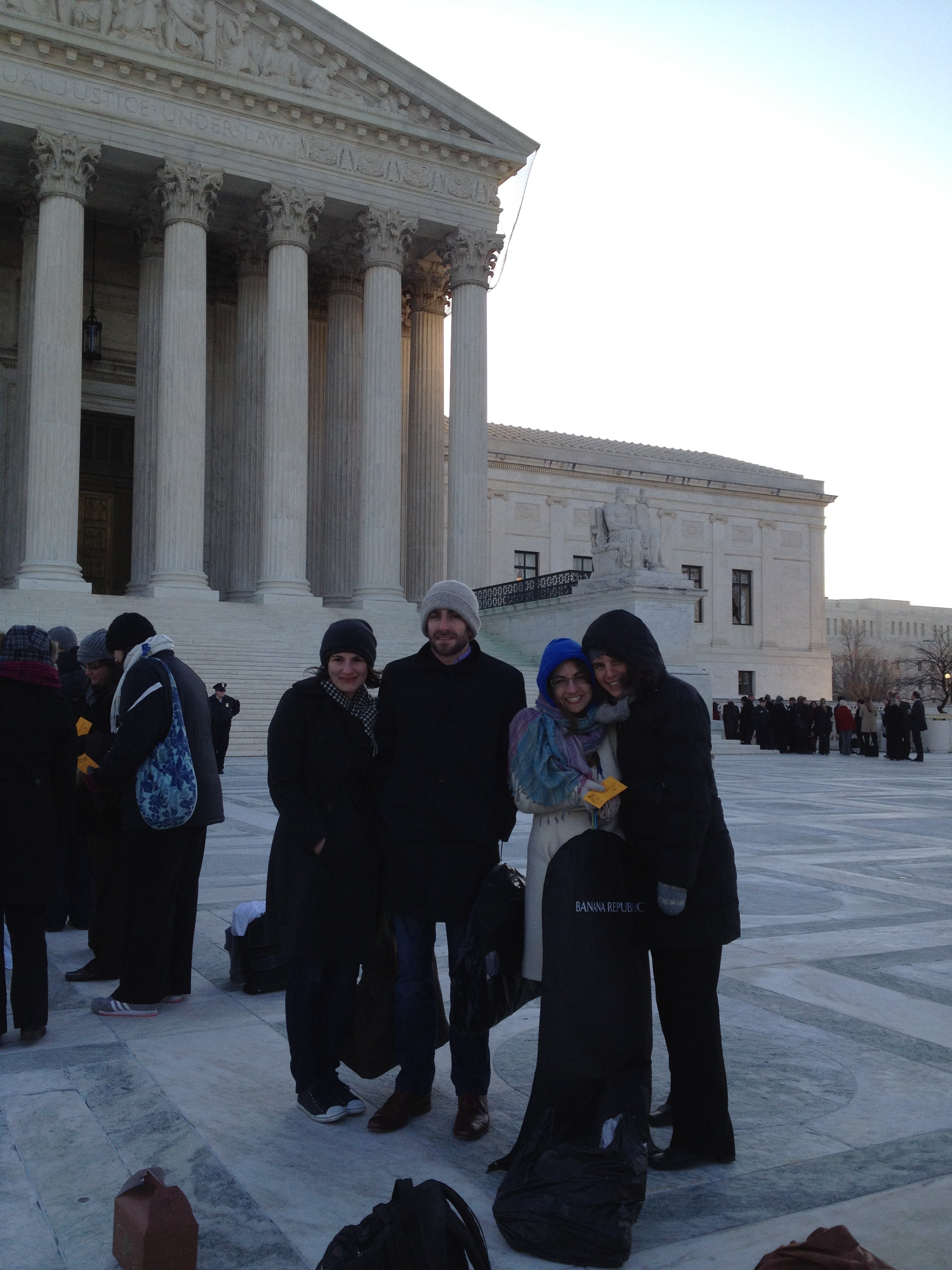 Four individuals smile outside the Supreme Court. They wear heavy coats, indicating it is winter.