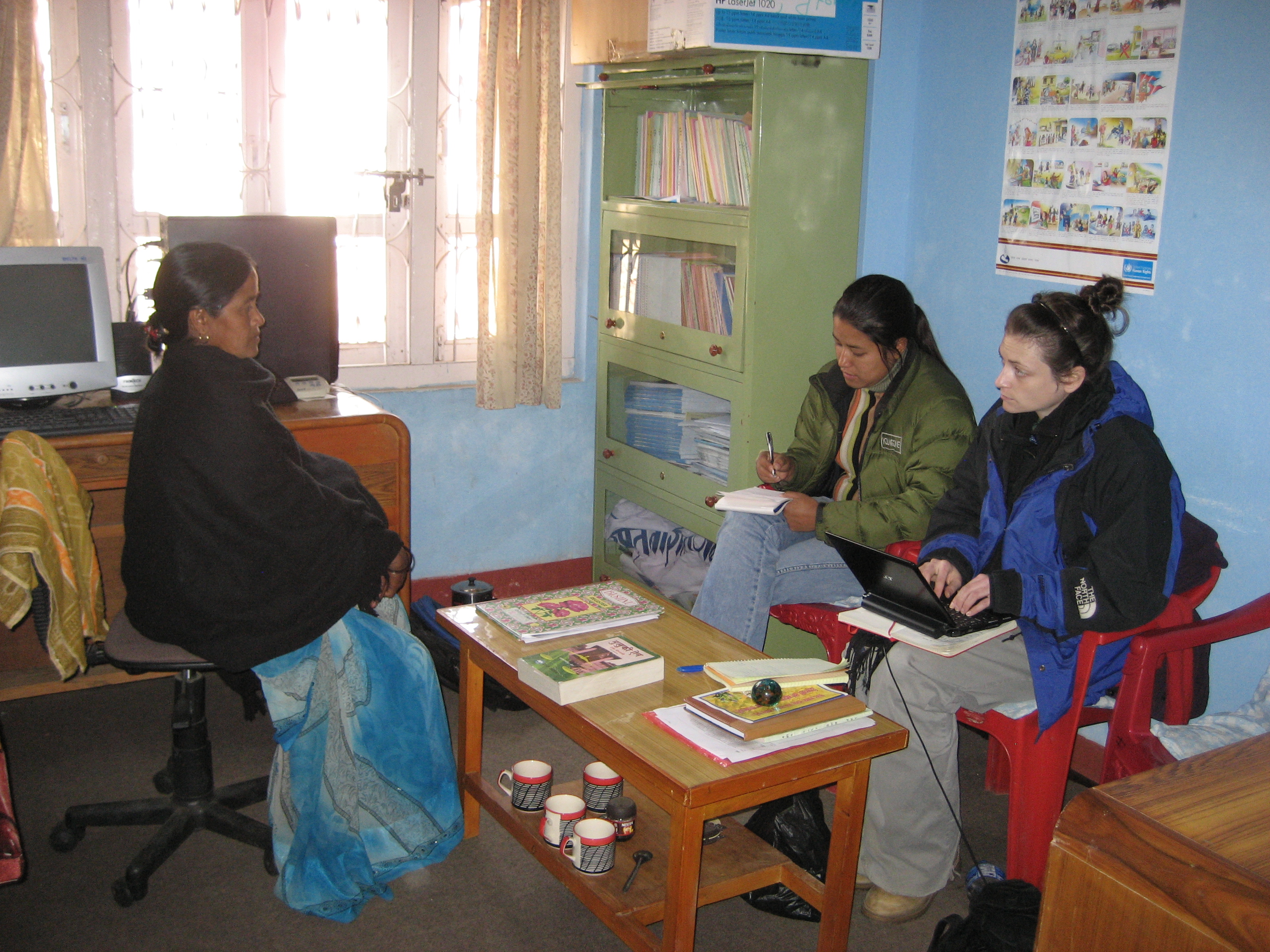 Two students wearing heavy coats interview another woman wearing a long skirt and fleece. There is a computer behind them, and they look to be a in an office setting.