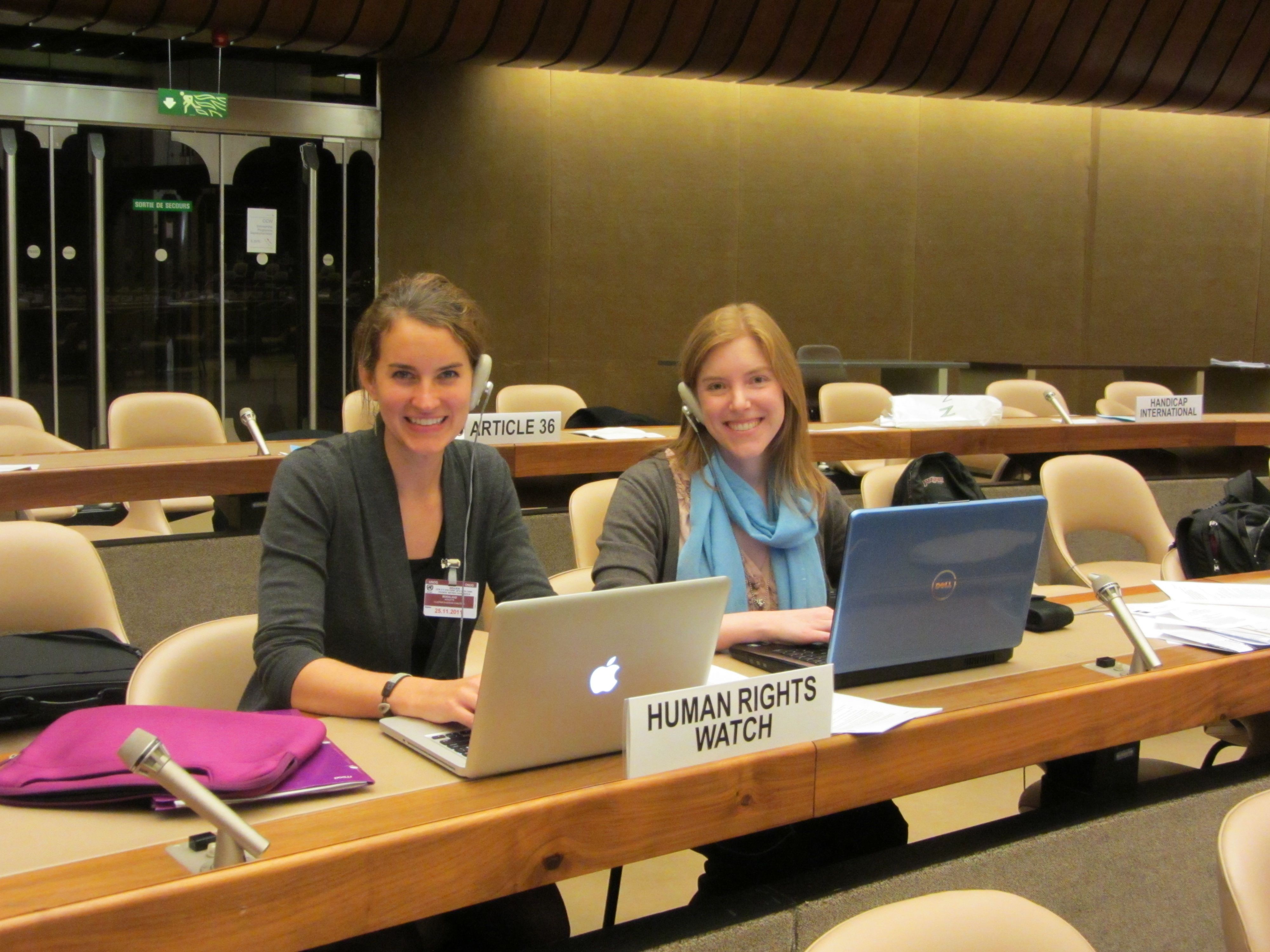Two women wear headsets in front of computers in a conference setting. A tag saying "Human Rights Watch" sits in front of them designating them as representatives of the NGO.