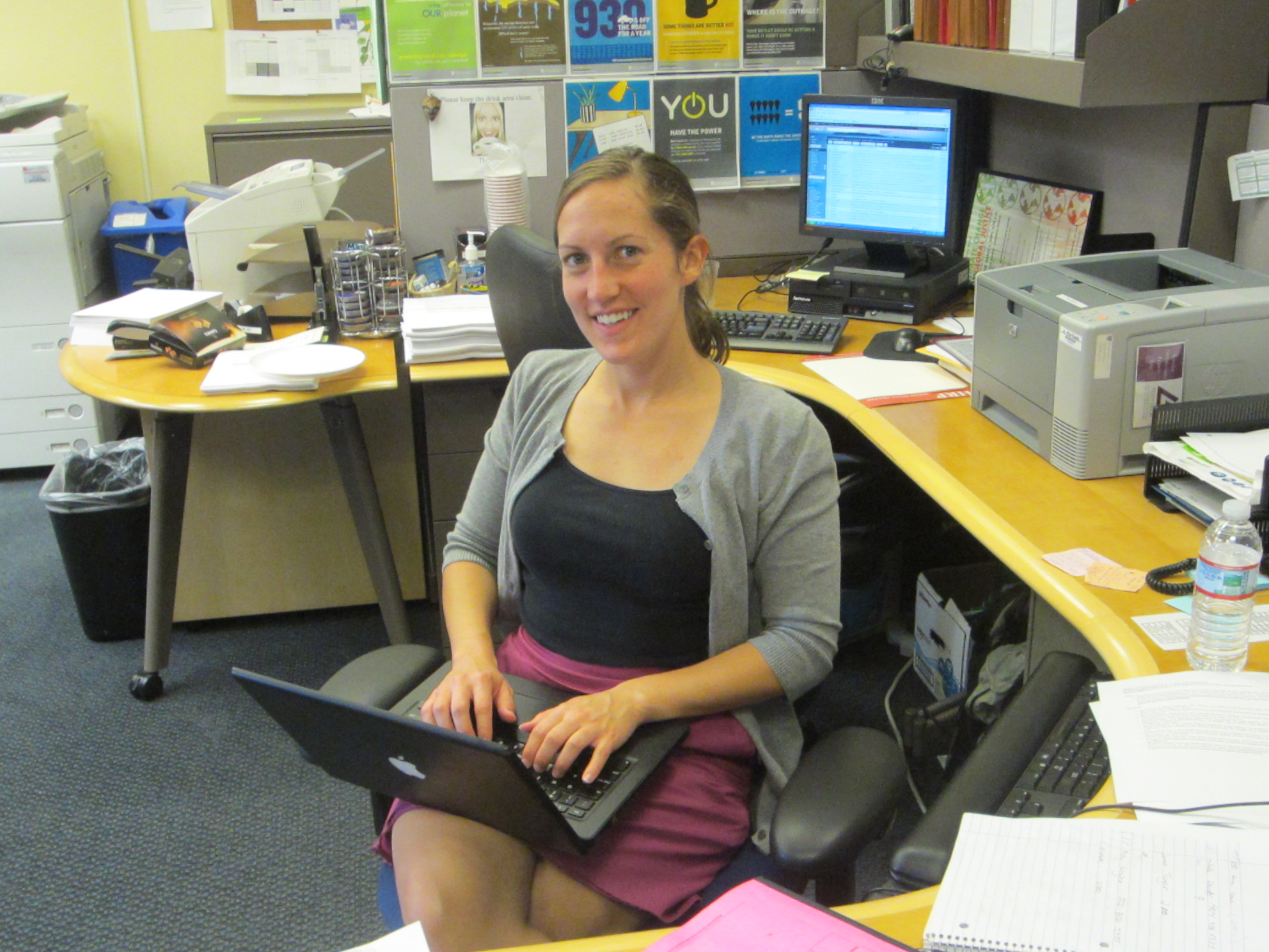 A woman wearing a sweater and a purple skirt sits at a cubicle with a mac laptop on her lap.