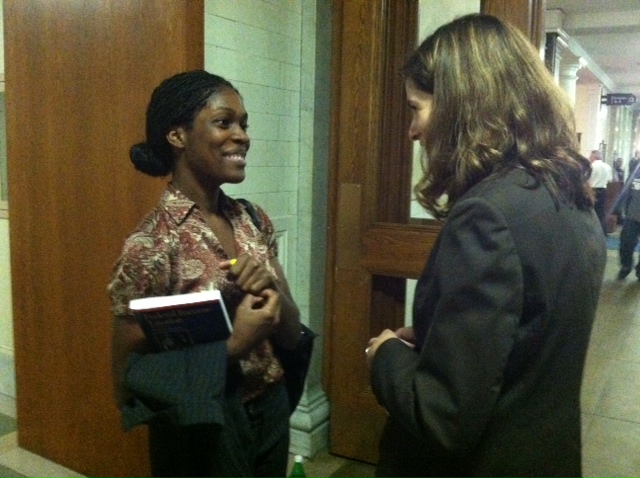 A woman holding a legal textbook speaks to another woman wearing a blazer. They are both smiling.