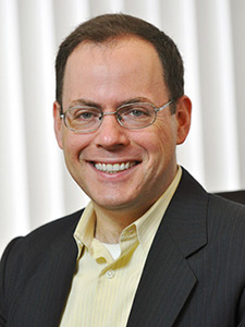Headshot of a man wearing glasses, a black blazer, and a yellow shirt.