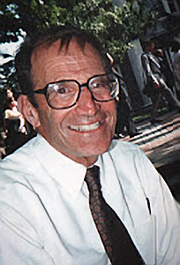 A man in his middle ages smiles at the camera. He wears a white button-down shirt and a tie and large square glasses.