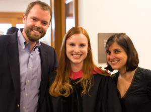 Two faculty smile with a student in graduation robes.
