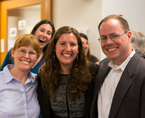 Three individuals smile for a photo while someone sneaks up behind them.