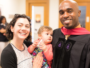 A man in a graduation robes smiles with a professor holding a baby.