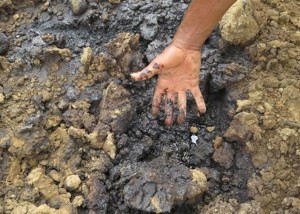A hand reaches down into soil that looks contaminated by oil.
