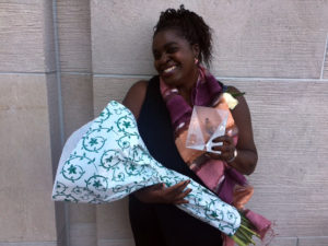 A woman holds flowers and an award and smiles against a building.