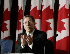 A man wearing a suit and tie gestures in front of five Canadian flags.