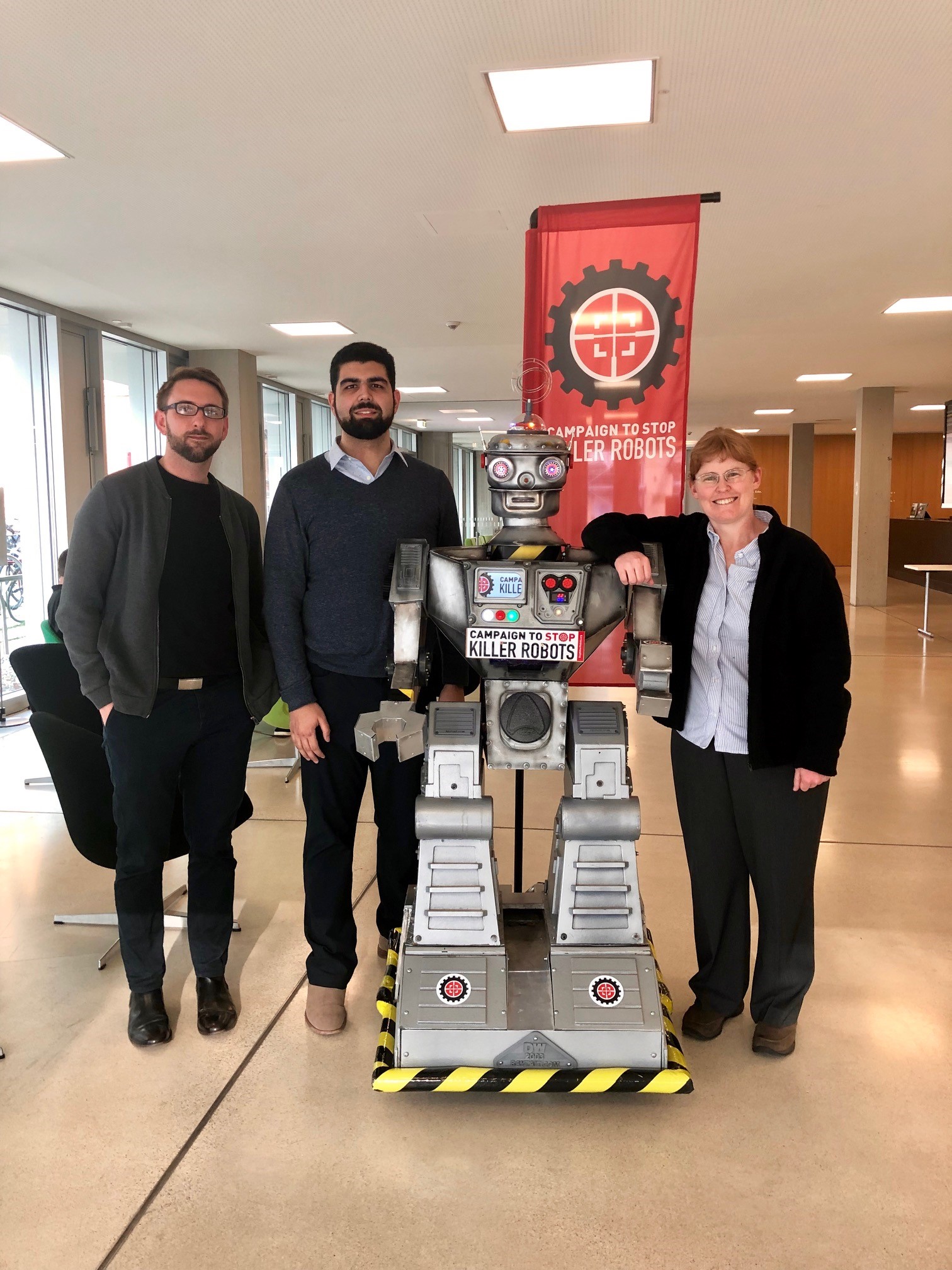 Three individuals pose with a life-size robot in front of a banner 