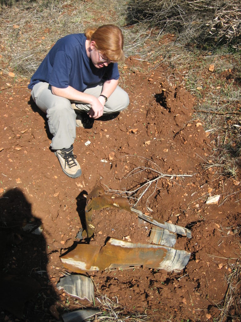 A woman kneals in the dirt examining the remainder of a cluster munition. It looks like broken stone buried in mud.