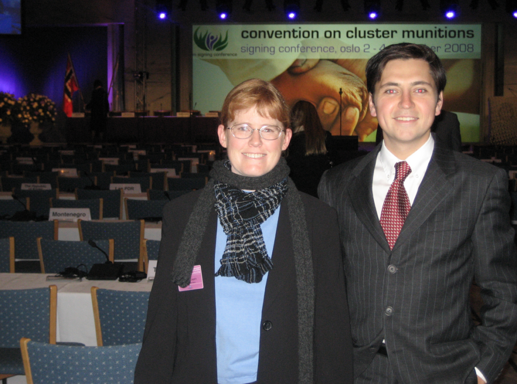 Bonnie, a woman with red hair, stands next to her student, Chris, both of whom wear formal wear. They stand in front of a banquet room with a slide behind them that notes "Convention on Cluster Munitions," and that it is the signing conference.