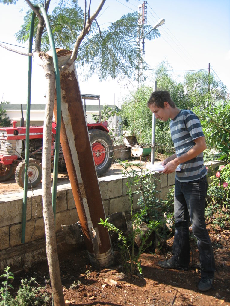 A young man examines a cluster munition shell all rusted out leaning up against a tree. He stands on red mud next to a wall. A red truck is in the background.