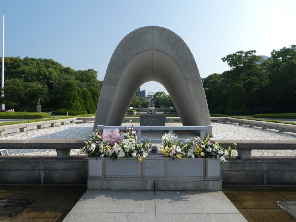 An arch with flowers in front of it honors those who lost their lives in Hiroshima on a clear day with trees behind it.