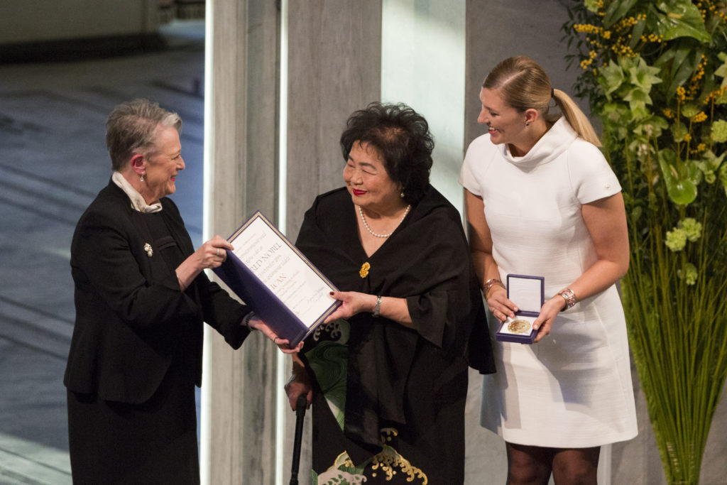 An elderly woman wearing a black drapey dress smiles as she is handed an embossed folder by another woman on the Nobel Prize Committee. Next to them, the Executive Director of ICAN, a tall blonde woman smiles and holds the Nobel Prize medal.