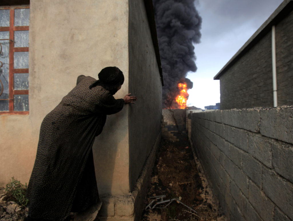A woman peers behind a wall as she sees smoke billowing and fire.