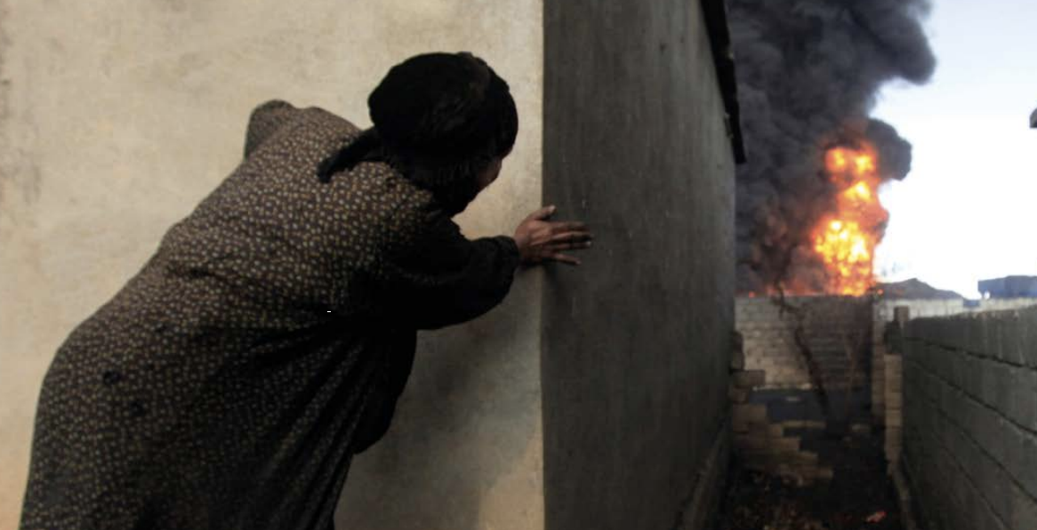 A woman hides from an oil fire in the distance.