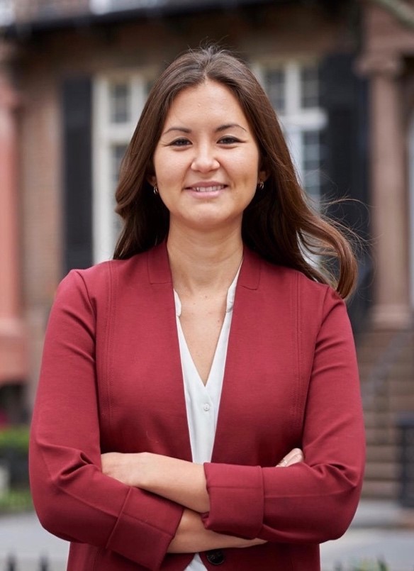 A woman with brown hair smiles in front of Harvard building. She wears a red blazer and a white shirt.