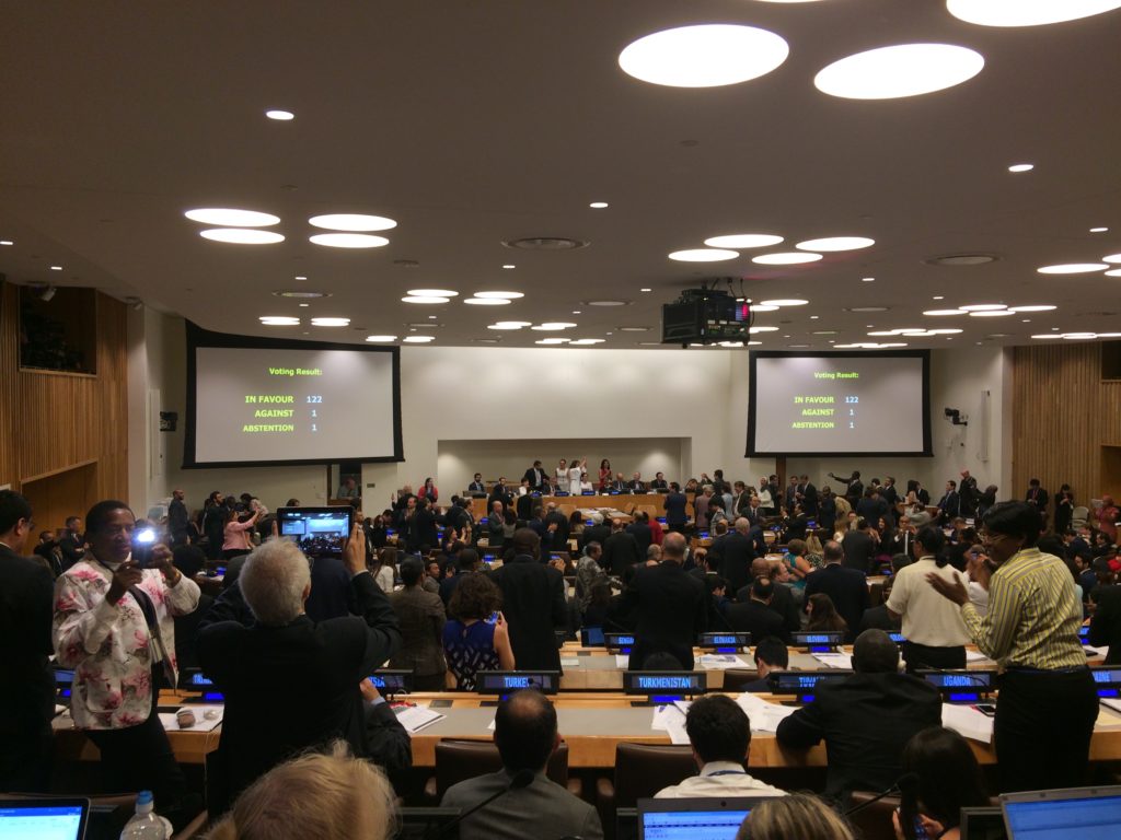 The backs of individual heads in a negotiation room look at screens that note 122 countries were "in favor" of the treaty.