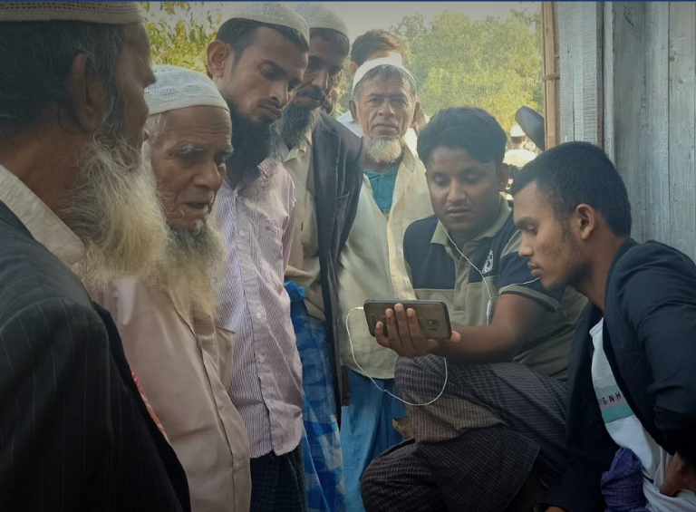 A group of Rohingya refugees stand in a circle, some with mouths agape as they crowd around a photo watching something.