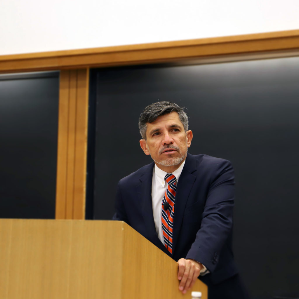 Victor Madrigal-Borloz stands in front of a podium in Wasserstein Hall. He wears a suit and tie and is speaking. He stands in front of a chalkboard.