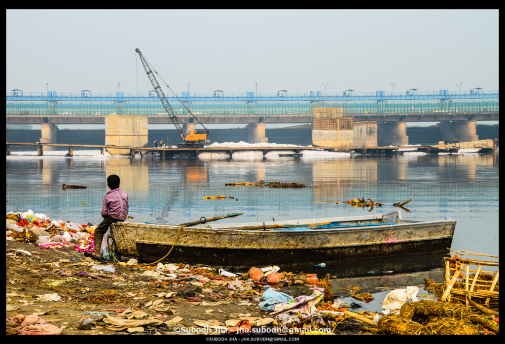 A young boy sits on a trash-ridden beach in front of the Yamuna River.
