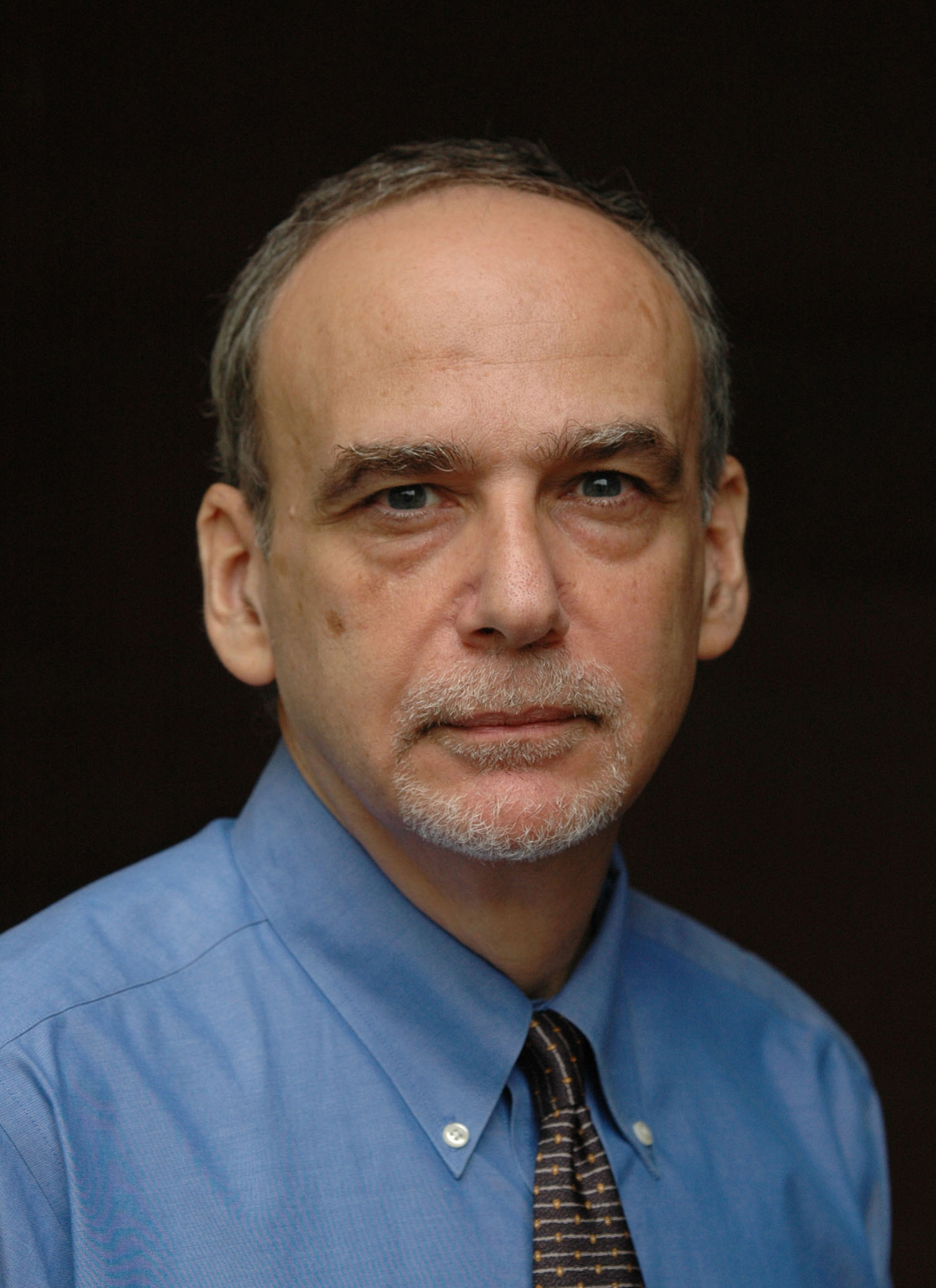 A man wearing a blue shirt and tie looks directly at the camera; behind him there is a black background.