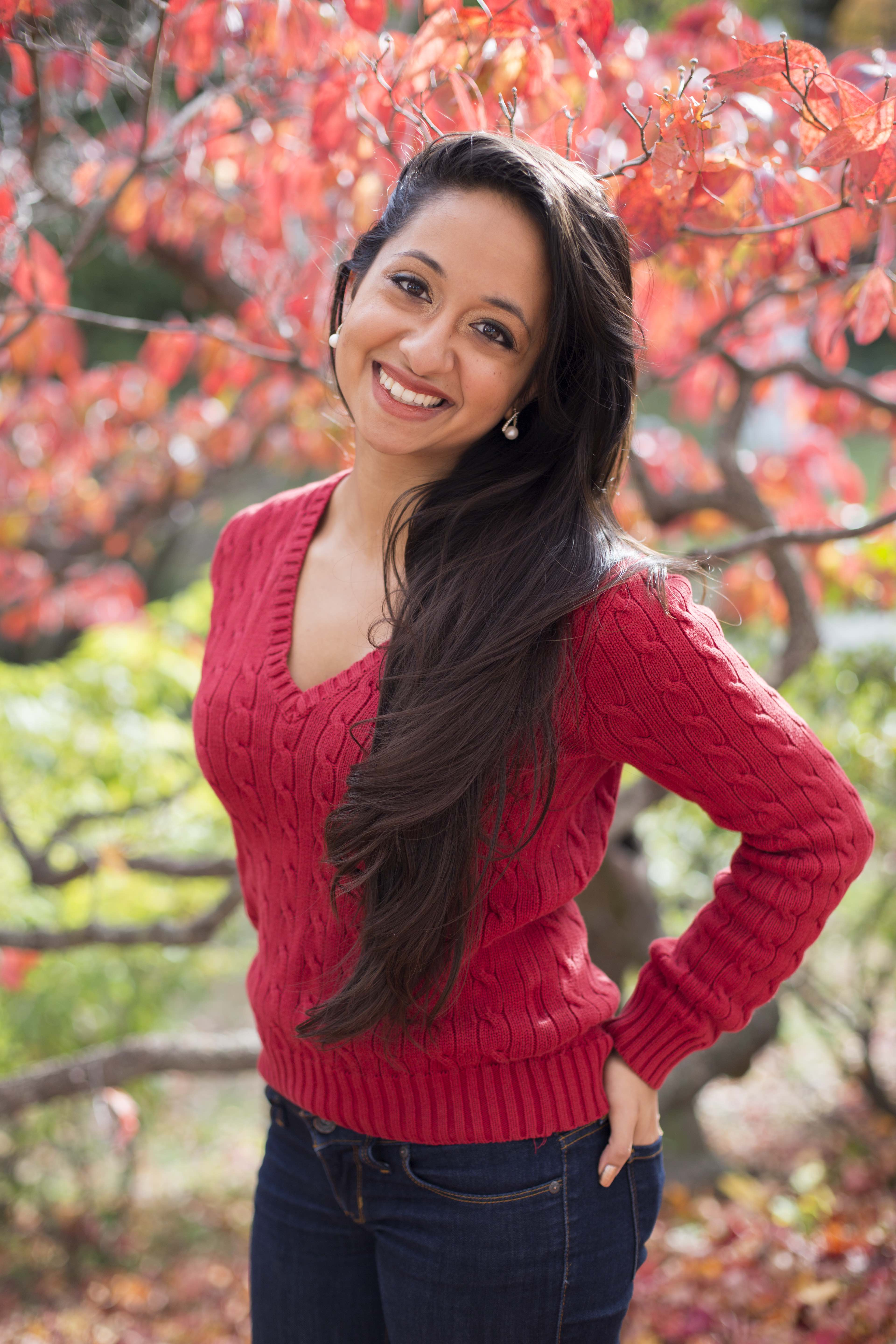 A young woman with a red sweater and jeans on smiles in front of the changing autumn trees.