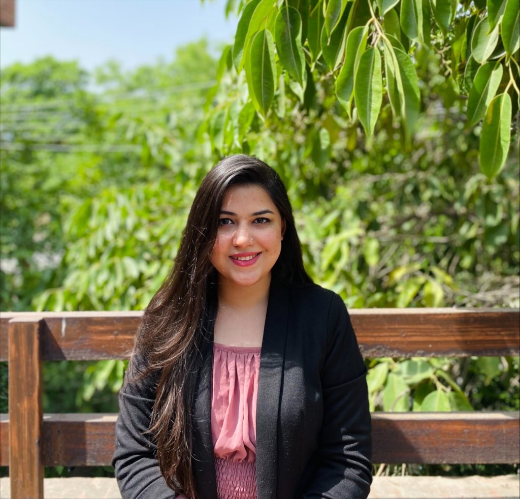Seher Aftab sitting on a wooden bench in front of lush green trees. 