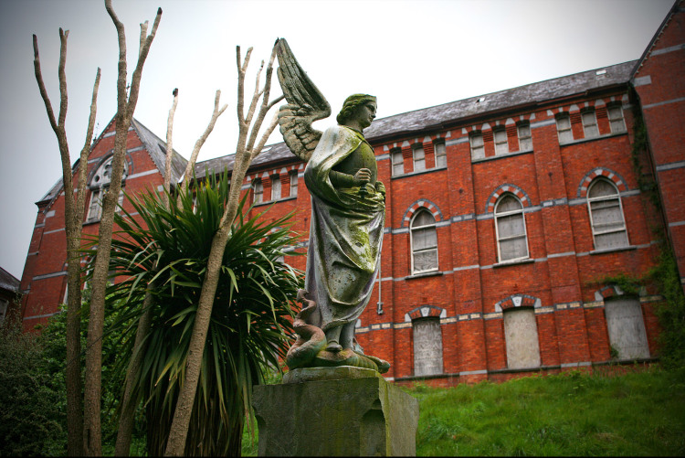 Angelic statue stands in-front of red brick Irish Magdalene asylum building.