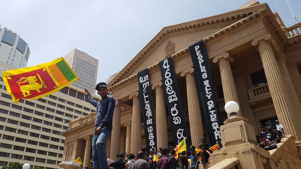 Protestor waving Sri Lankan flag in front of imposing government building. 
