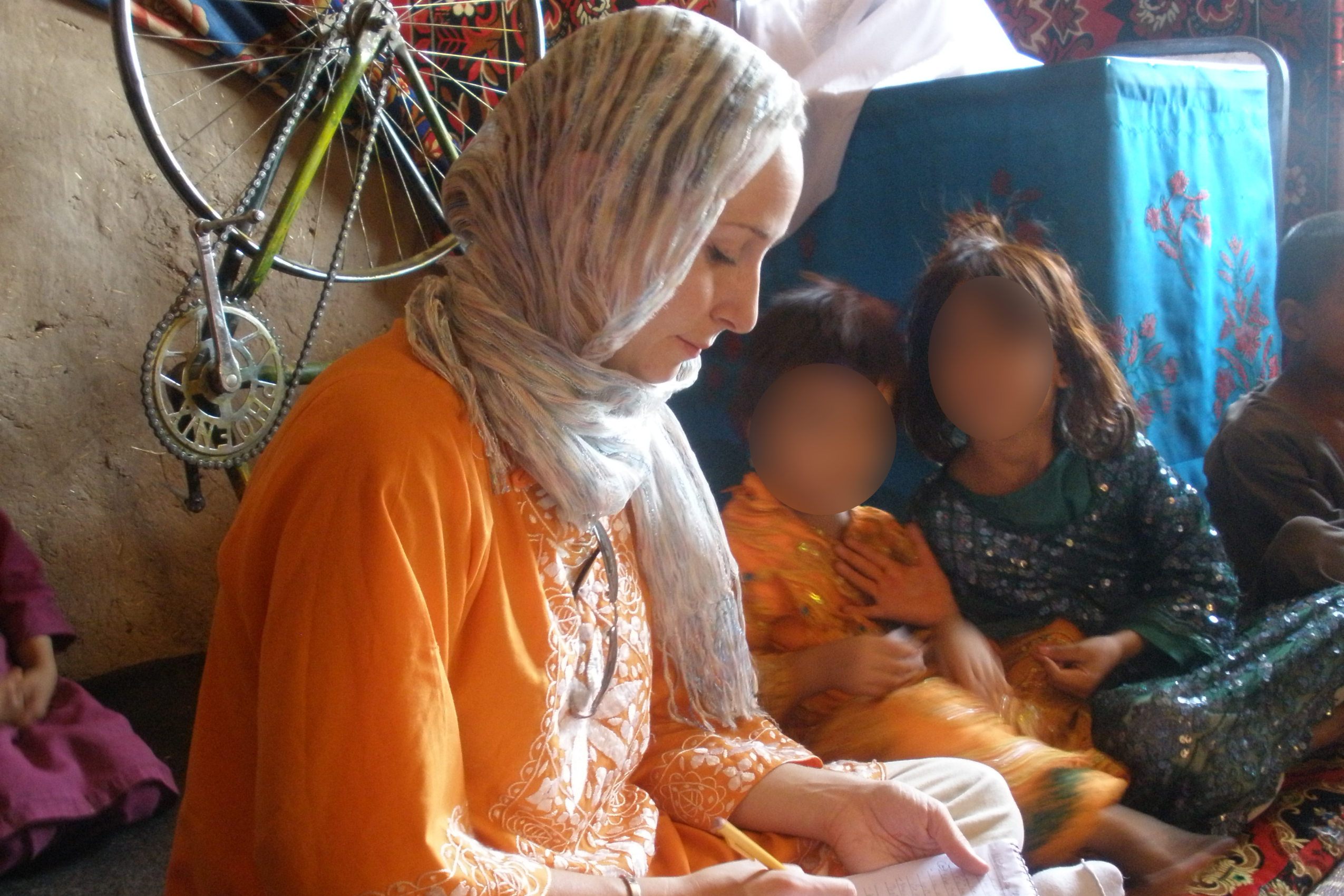 Erica Gaston jotting down notes while sitting cross-legged in a residence in Afghanistan. Children are in the background.