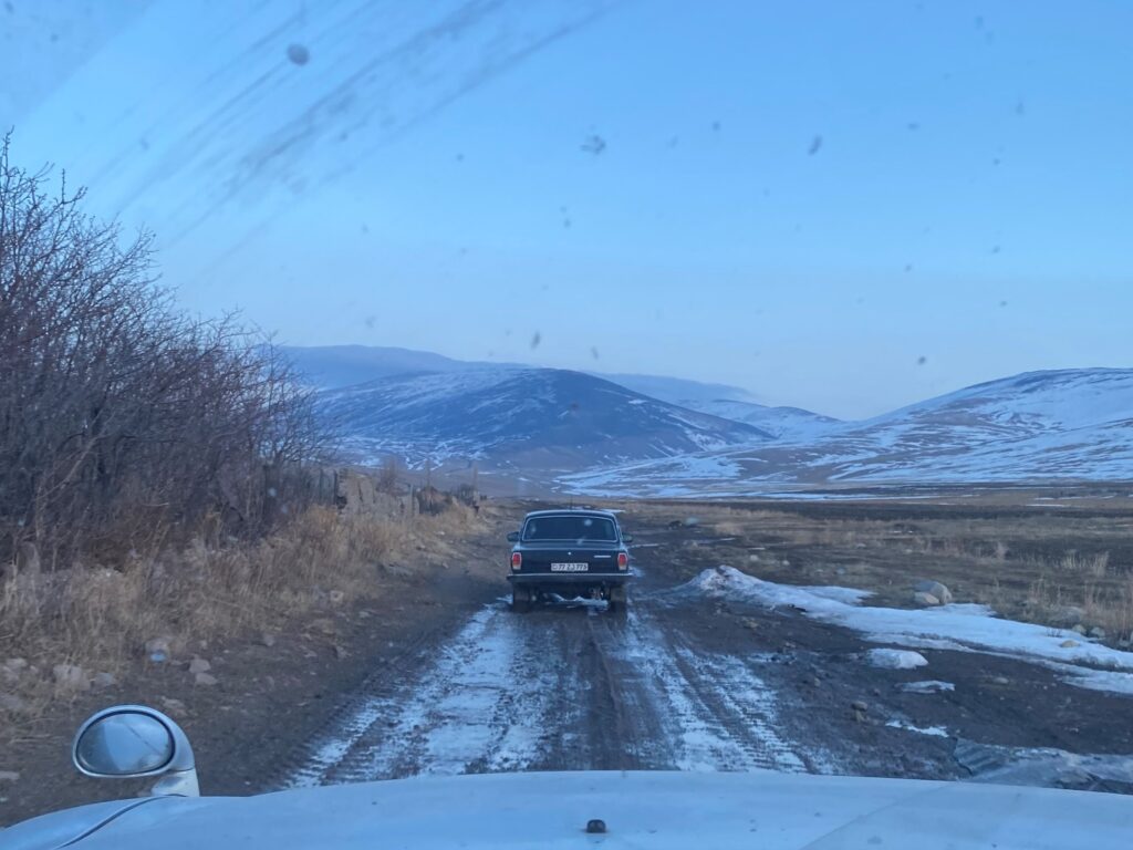 A photo of the back of an old car driving on a muddy road in front of snow-covered hills. The photo was taken from the front seat of a different car, and the hood is visible at the bottom. 