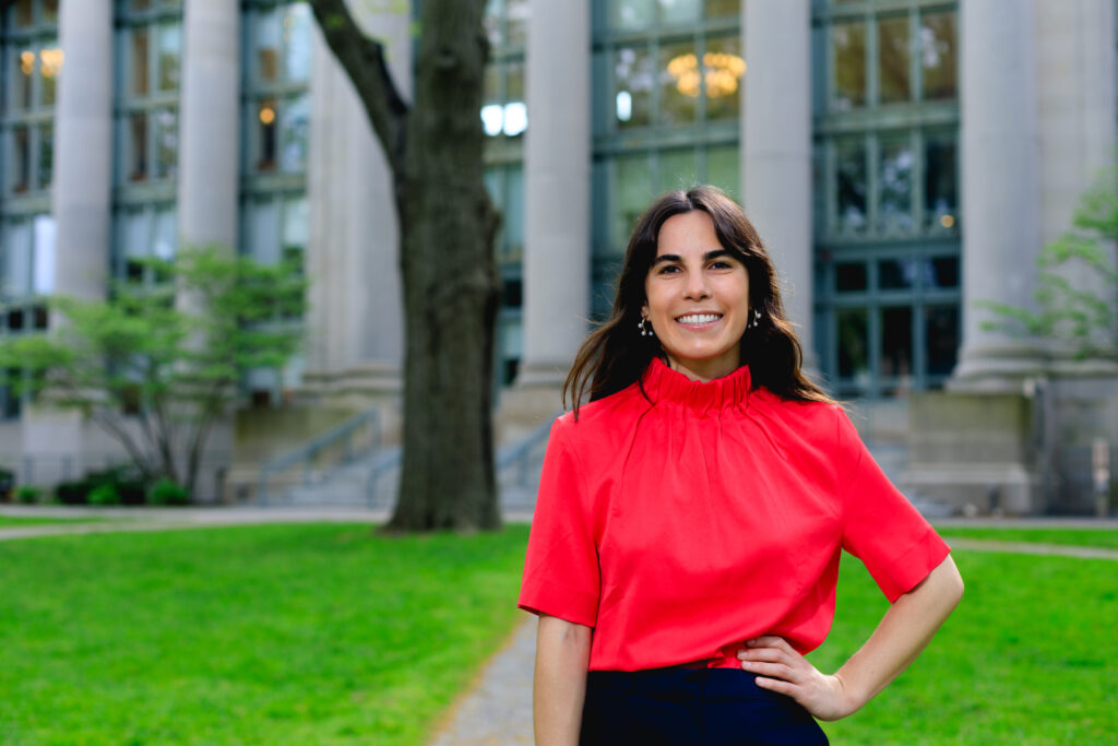 Rebecca Gore is wearing a red top and navy pants and is smiling into the camera. She is standing in front of the Harvard Law School Library.