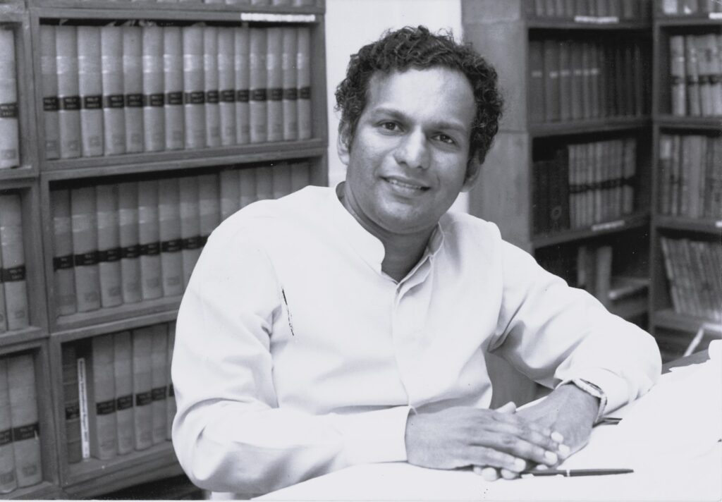 Black and white photo of Neelan Tiruchelvam sitting at a desk in front of book shelves smiling at the camera. 