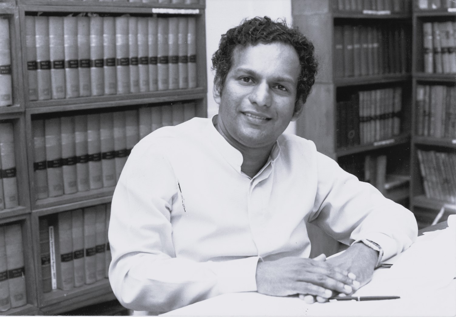 Neelan Tiruchelvam sitting at table in front of book shelves and smiling into camera.