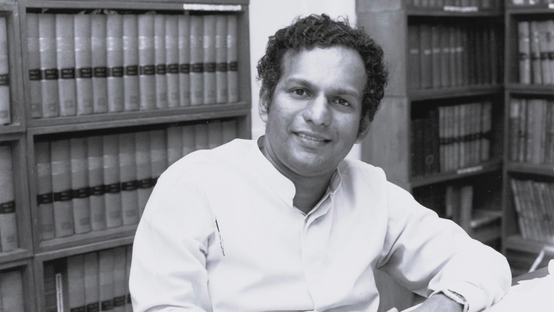 Black and white photo of Neelan Tiruchelvam sitting at a desk in front of book shelves smiling at the camera.