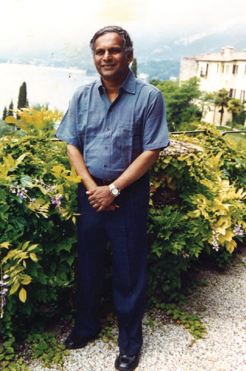 Neelan Tiruchelvam smiling dressed in a cerulean shirt and navy pants while standing in front of hedges. 