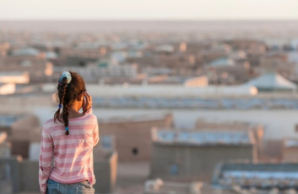 A young girl with braided hair, wearing a pink and white striped long-sleeve shirt and blue jeans, stands with her back to the camera while looking out over a vast, sunlit settlement of low, flat-roofed buildings that stretch into the distance. These buildings make up the Smara refugee camp.