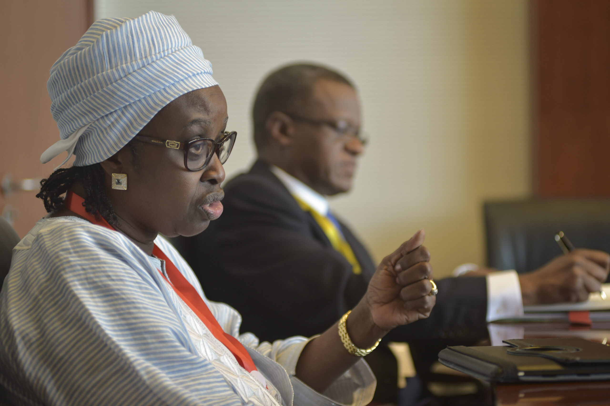 Woman in a striped headwrap and glasses speaks and gestures with her hand during a meeting, while a man in a suit sits slightly out of focus beside her taking notes at a conference table.