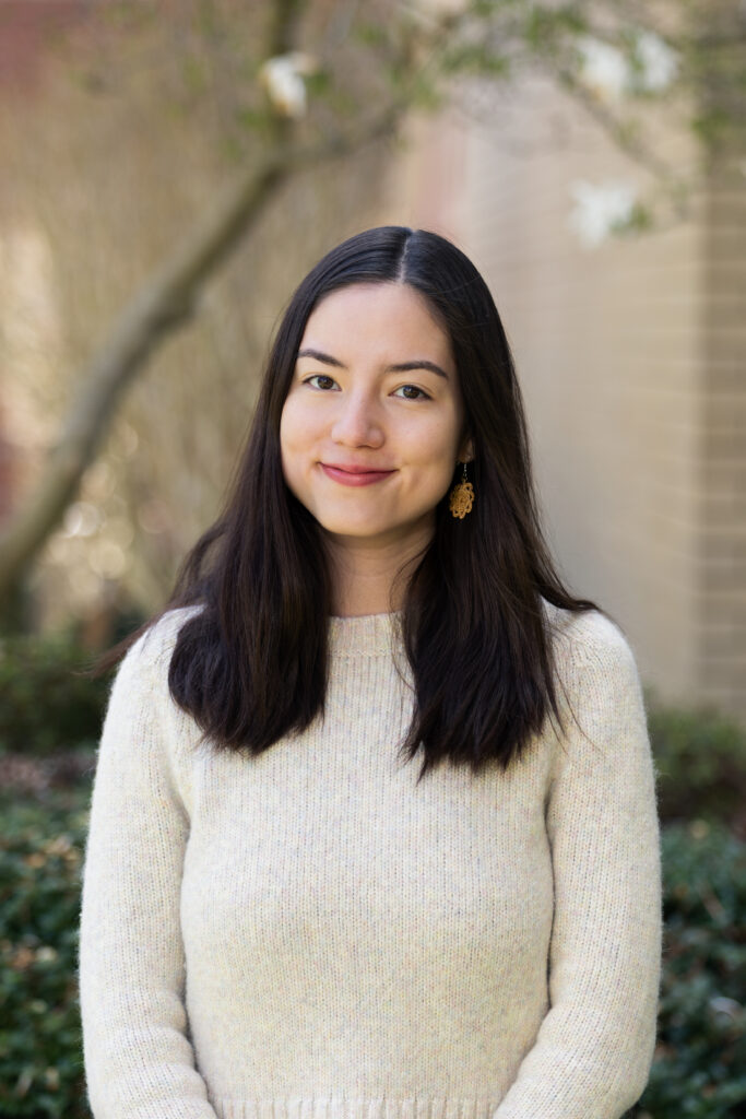 Headshot of a woman in a white sweater standing by a tree.